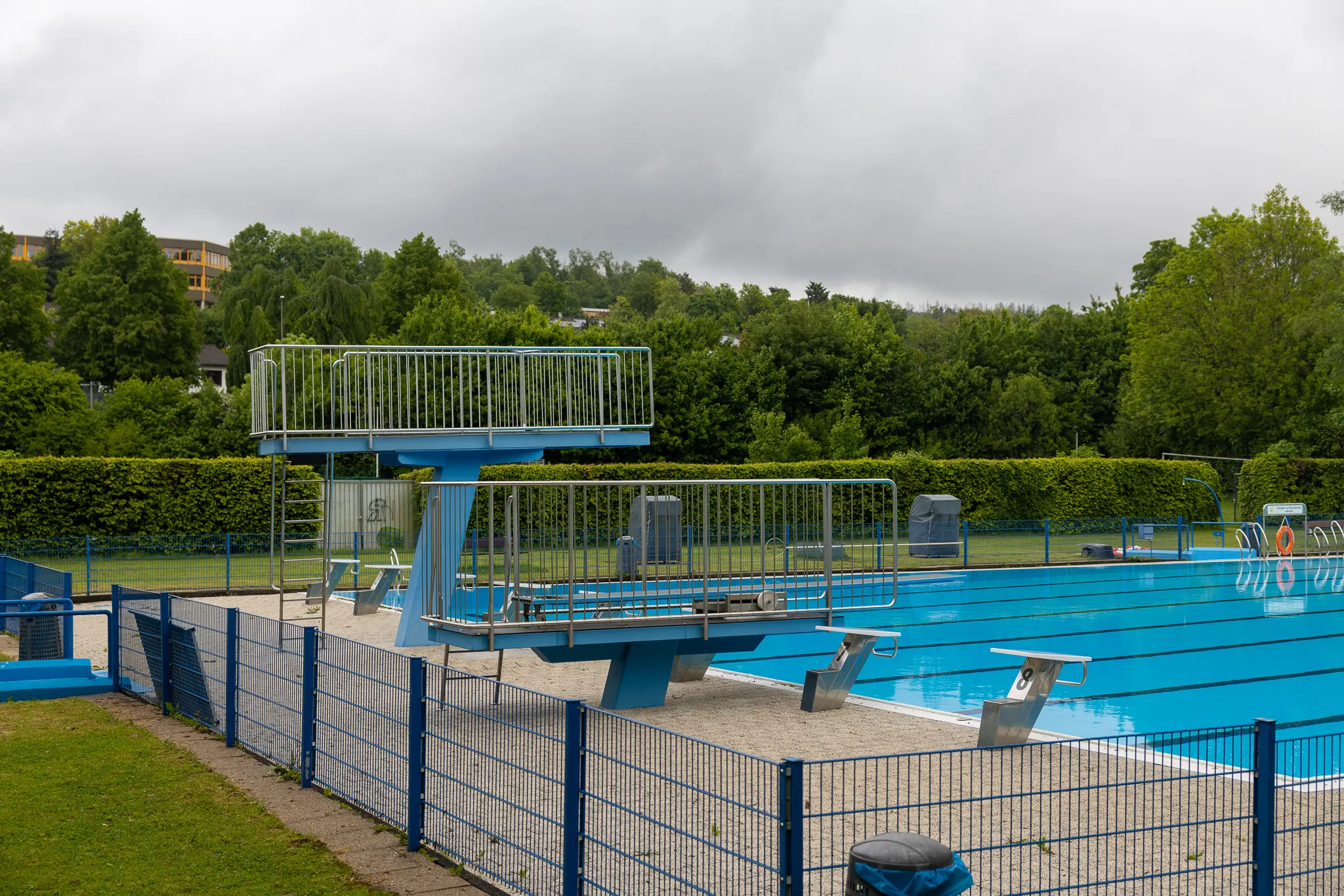 Blaues Sprungbrett vor einem Schwimmbecken mit einem Zaun davor, im Hintergrund Bäume und Gebäude unter einem bewölkten Himmel.