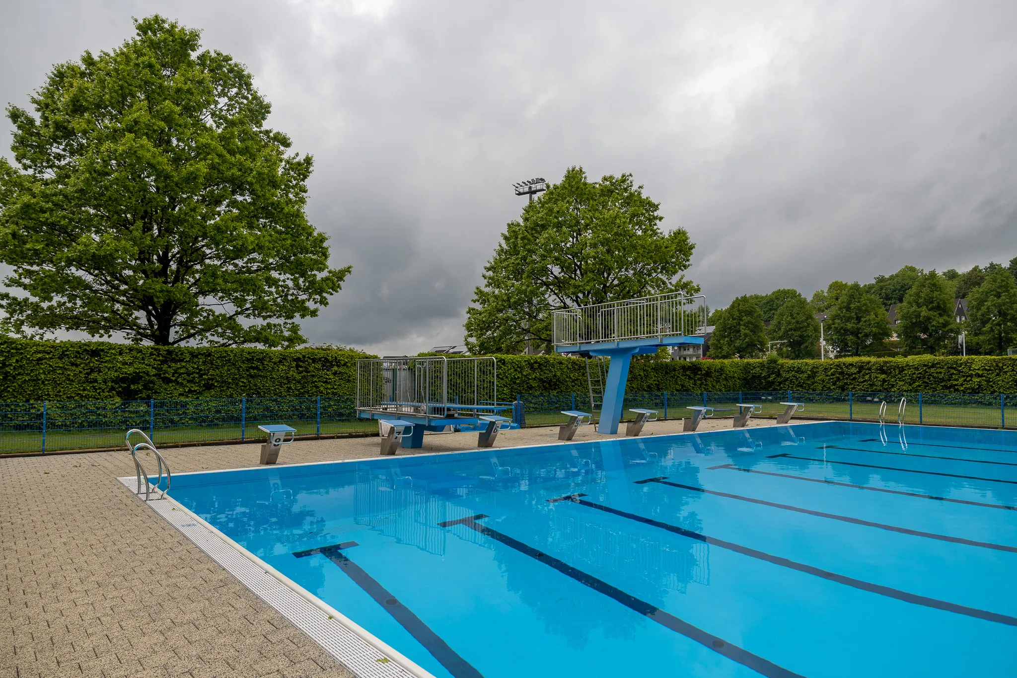 Schwimmbecken mit Startblöcken und Leitern, umgeben von Bäumen und einer Hecke unter einem bewölkten Himmel.