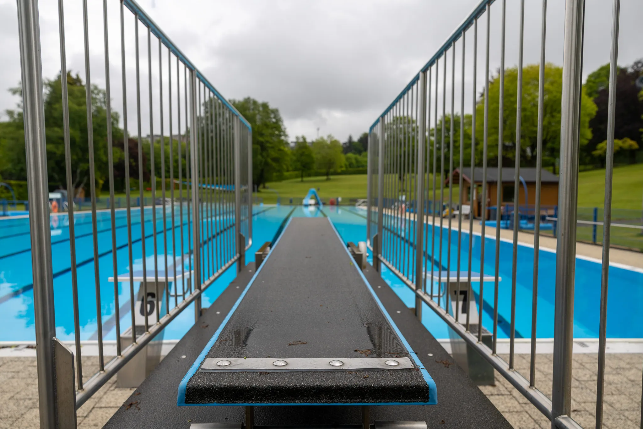 Blick vom Sprungturm über ein Schwimmbecken mit bläulichem Wasser und Bahnenmarkierungen, umgeben von Bäumen und Gebäuden.