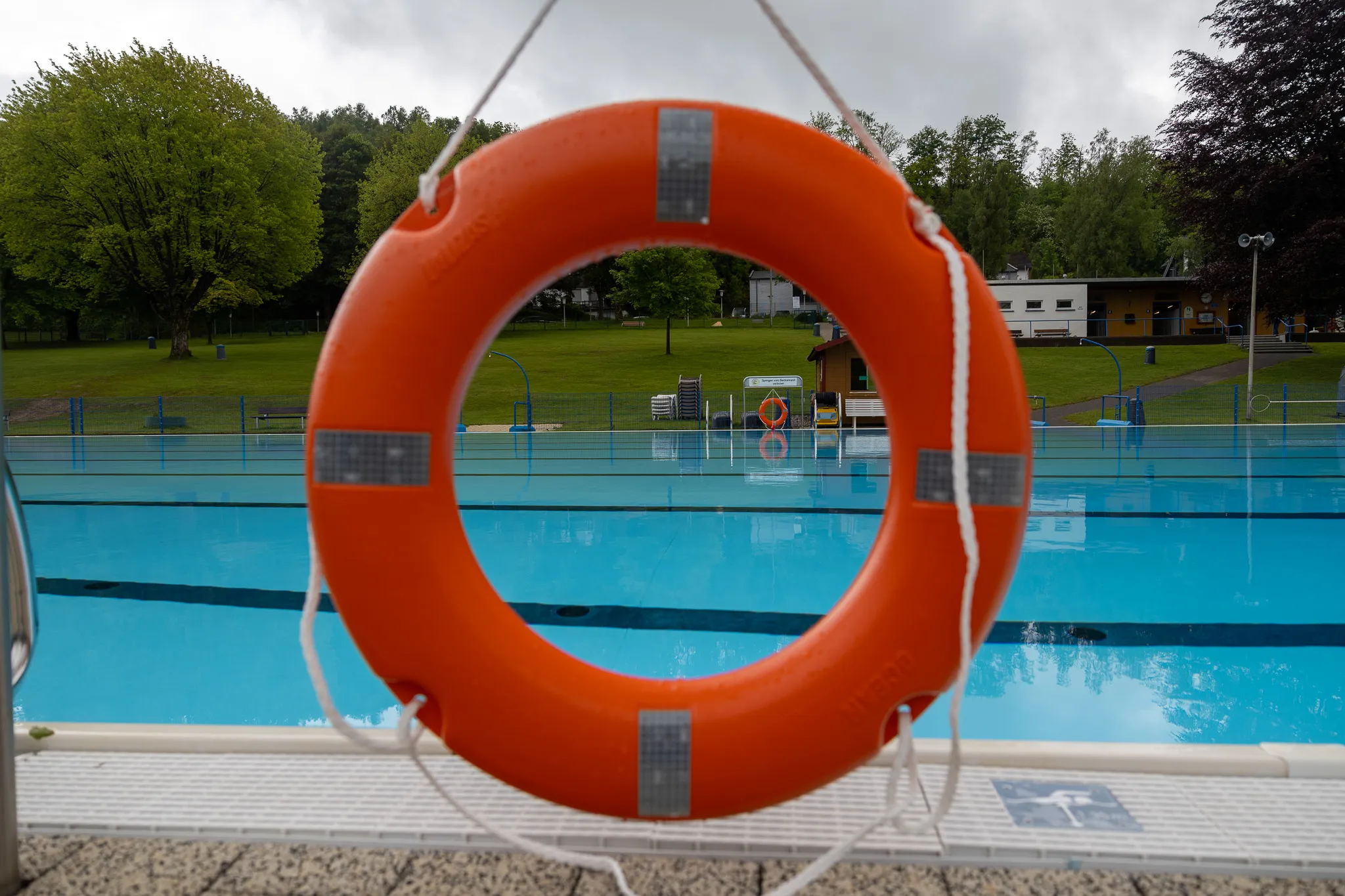 Ein orangefarbener Rettungsring hängt vor dem Becken eines Freibads, im Hintergrund Bäume und Gebäude.