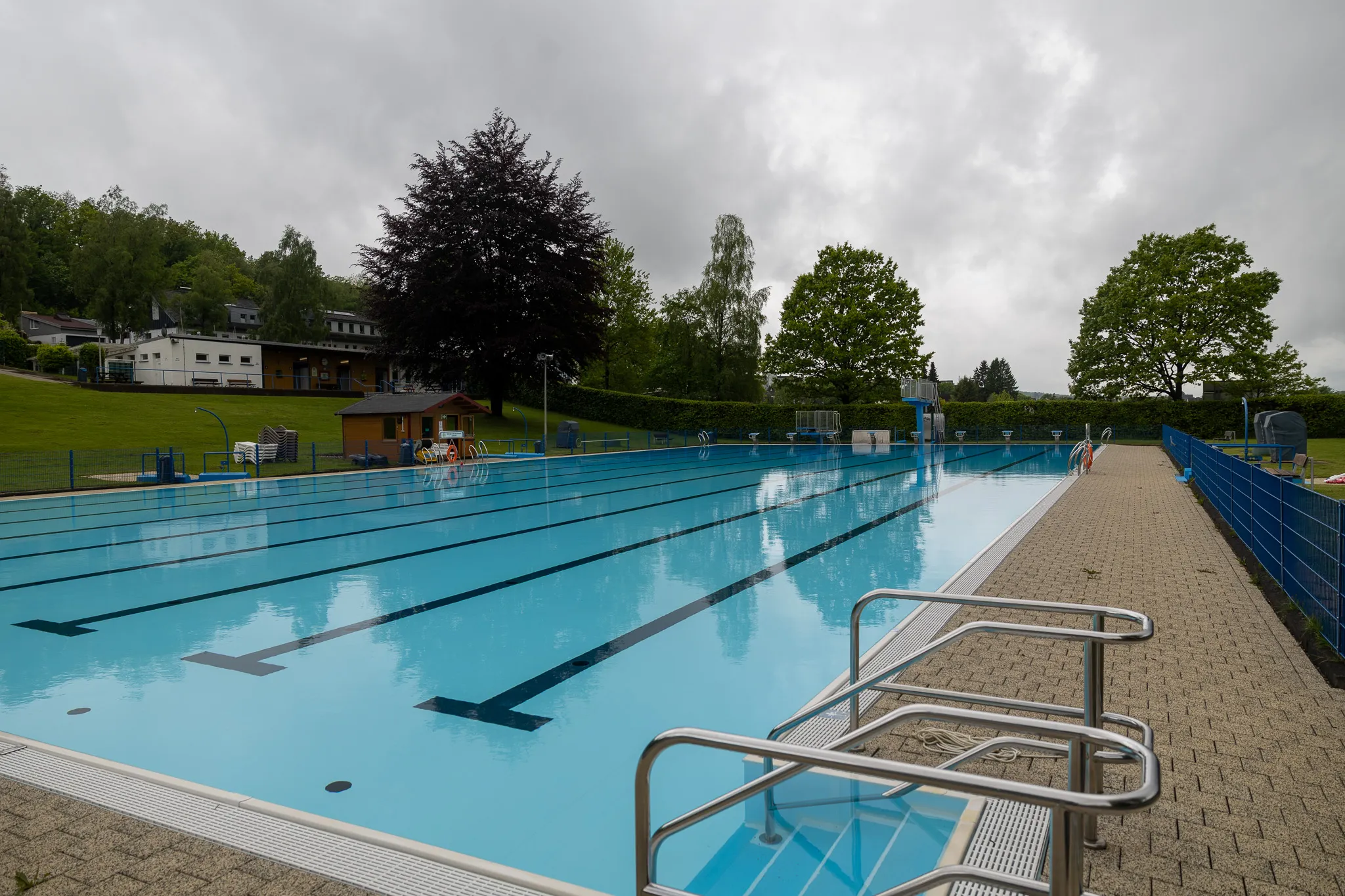 Leeres Schwimmbecken mit bläulichem Wasser an einem bewölkten Tag, umgeben von einem gepflasterten Bereich und Liegestühlen. Im Hintergrund ein Gebäude und Bäume.
