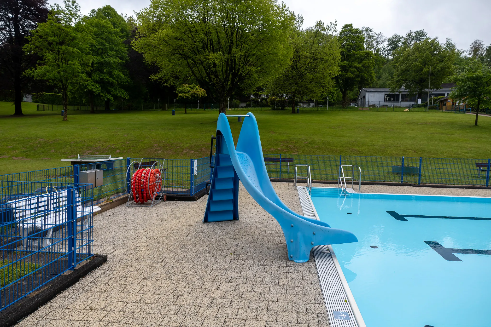 Blaues Schwimmbecken mit einer Rutsche und einer Leiter am Beckenrand, umgeben von einem gepflasterten Bereich und einer Grünfläche mit Bäumen.