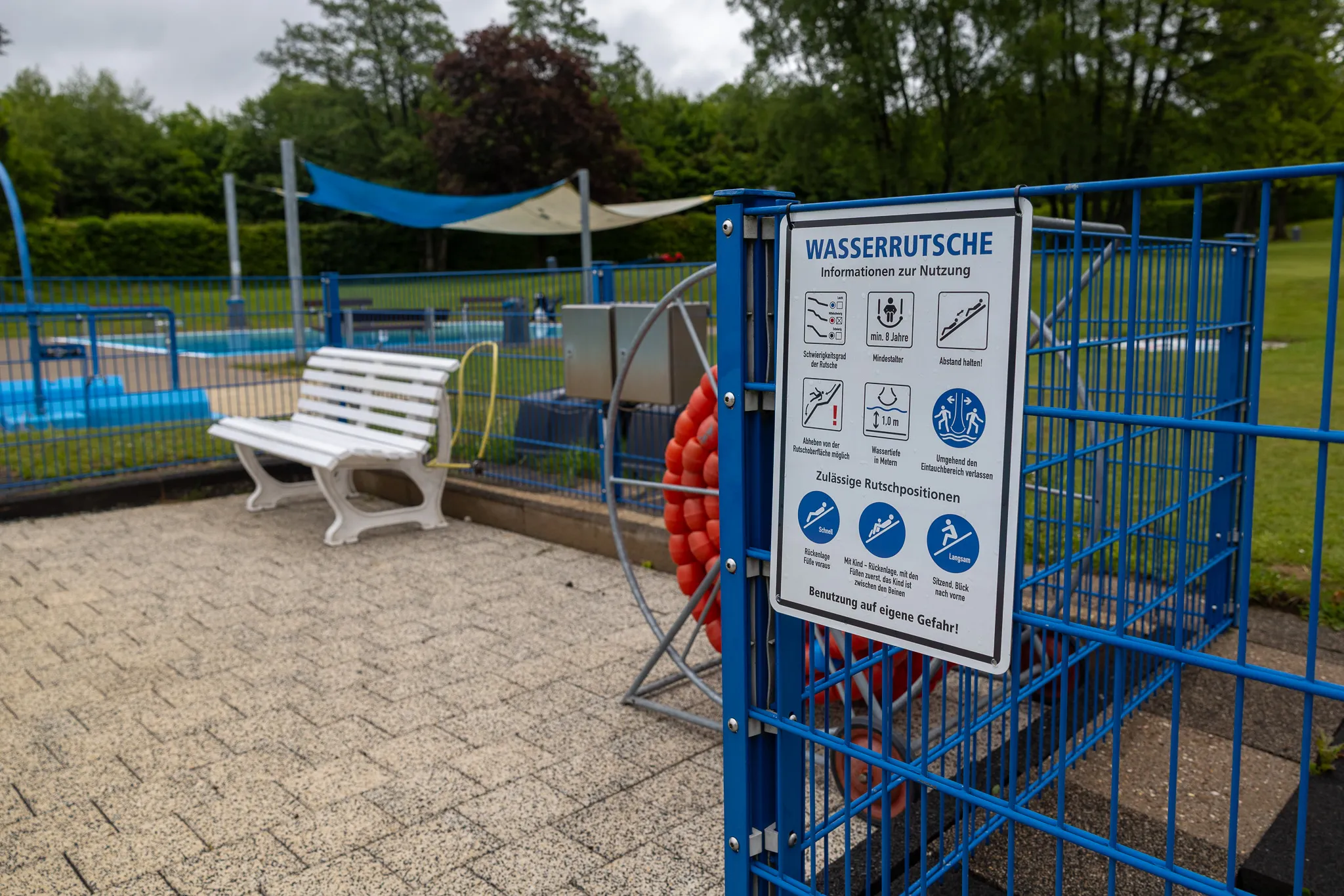 Schild mit Nutzungshinweisen für eine Wasserrutsche vor einem blauen Schwimmbecken im Freibad.