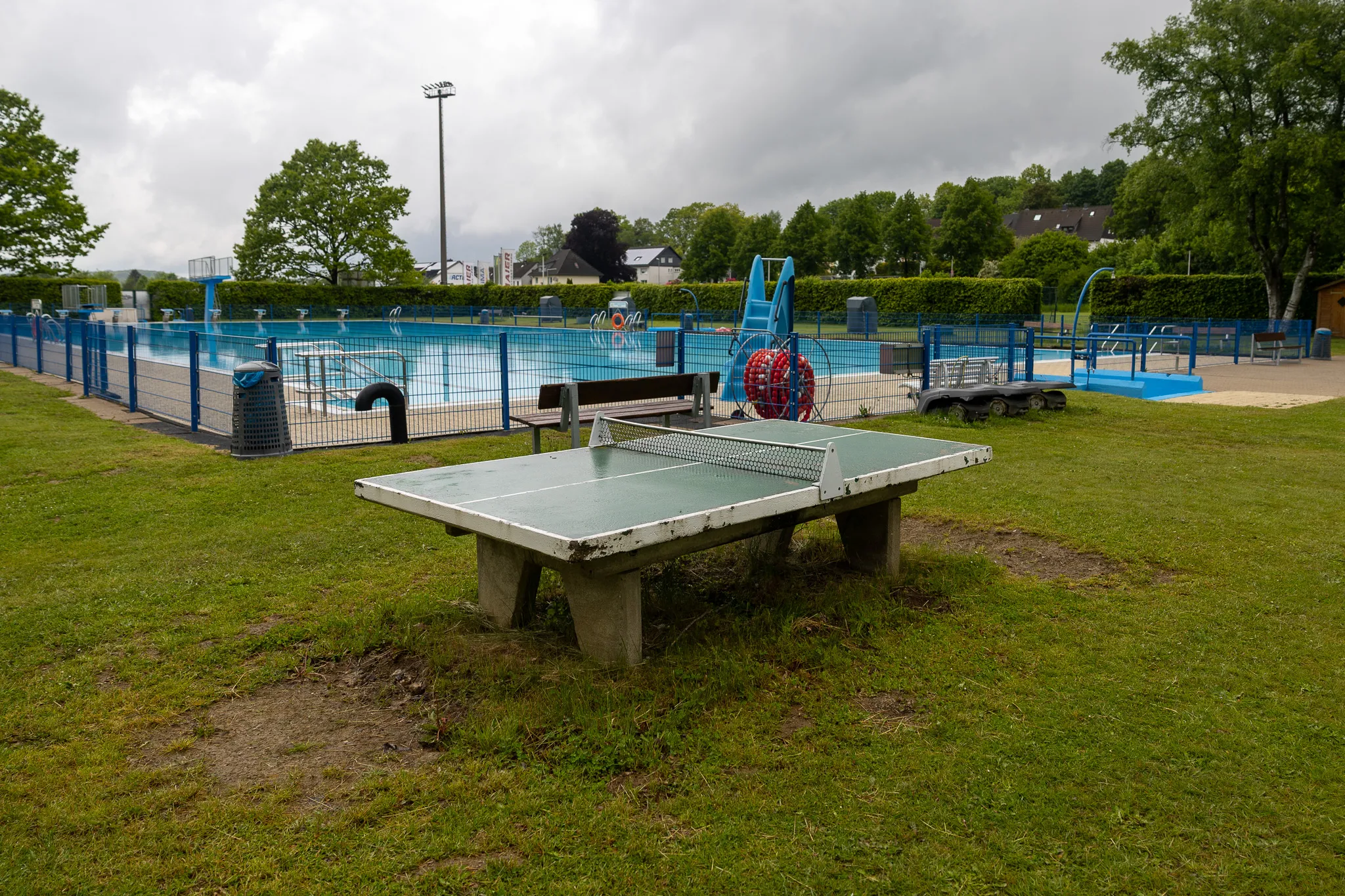 Tischtennisplatte auf einer Rasenfläche vor einem Freibad mit blauem Wasser und Bäumen im Hintergrund.