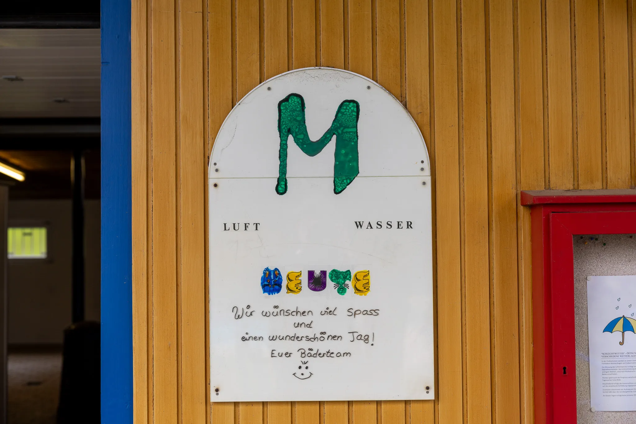 Schild mit dem Buchstaben M und den Wörtern „Luft“ und „Wasser“, darunter eine bunte Illustration und der Schriftzug „Wir wünschen viel Spaß und einen wunderschönen Tag!“. 