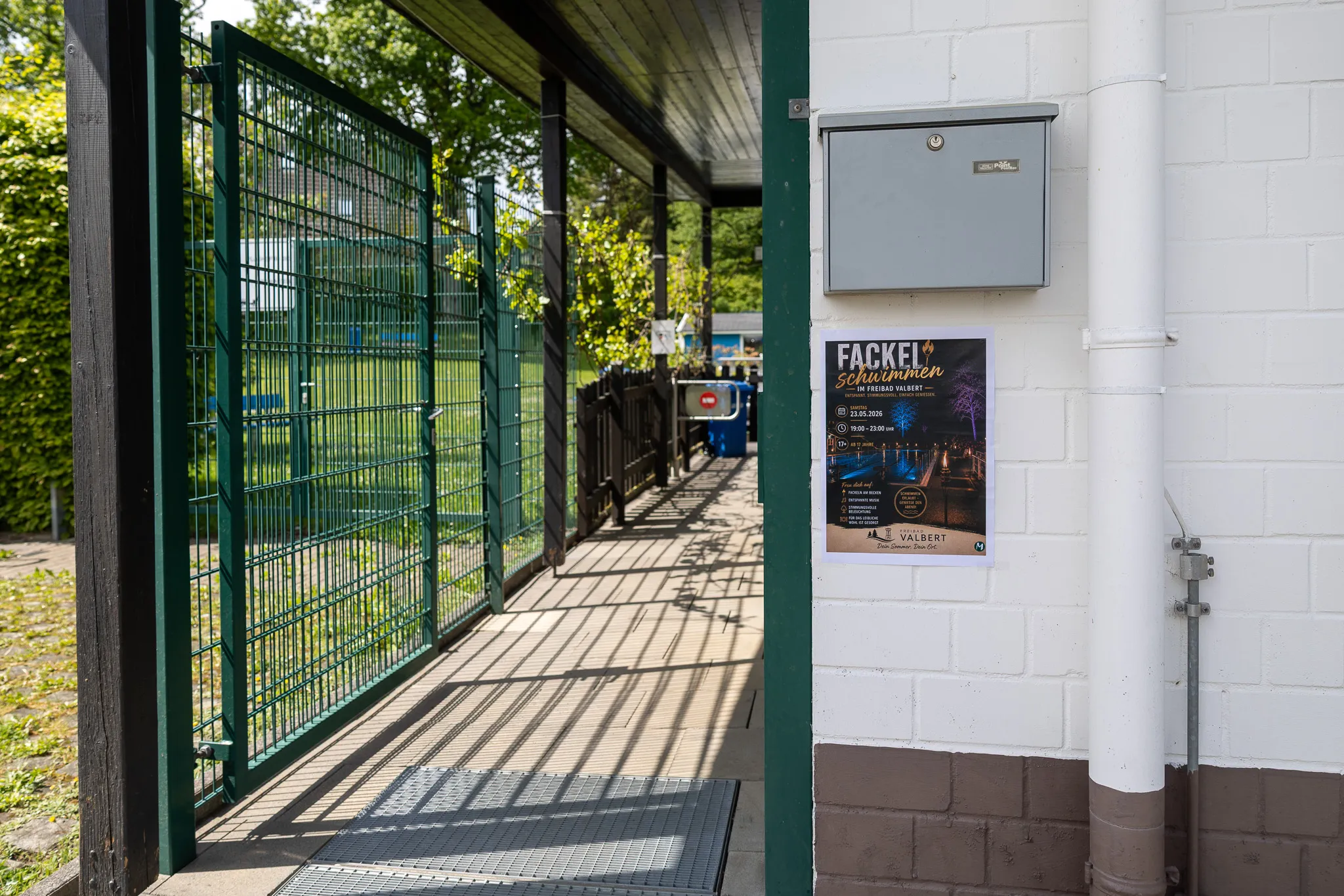 Ein Tor führt zu einem Pfad in Richtung eines Freibads mit grünem Gestrüpp im Hintergrund und einem Plakat mit Informationen zur Eröffnungssaison.