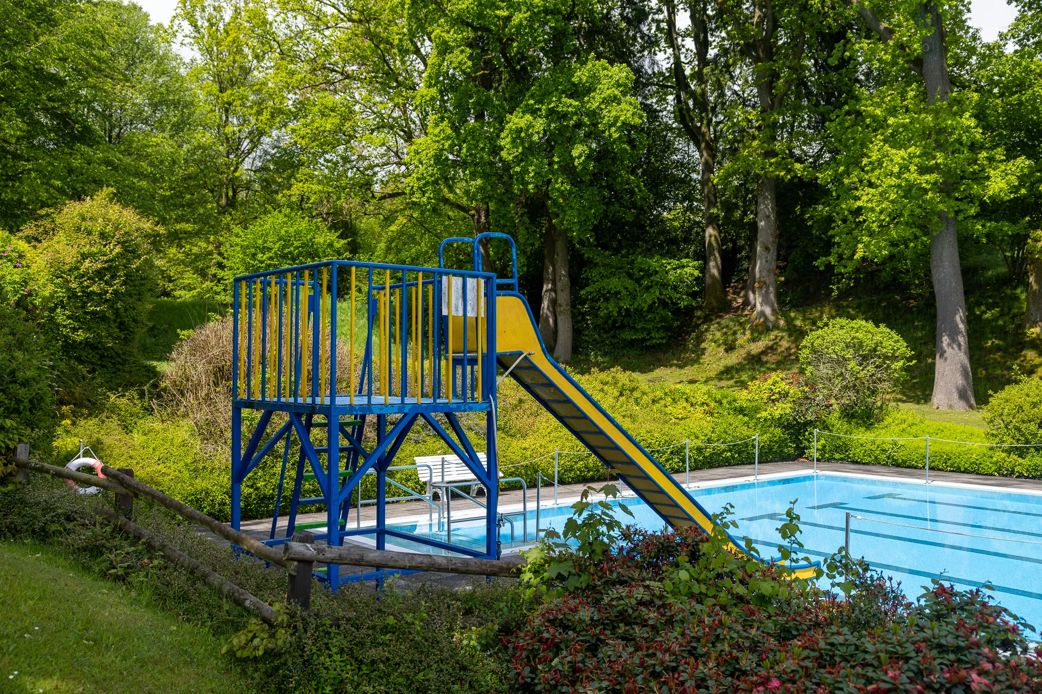 Blau-gelbe Wasserrutsche neben einem Schwimmbecken im Freien, umgeben von Bäumen und Büschen.