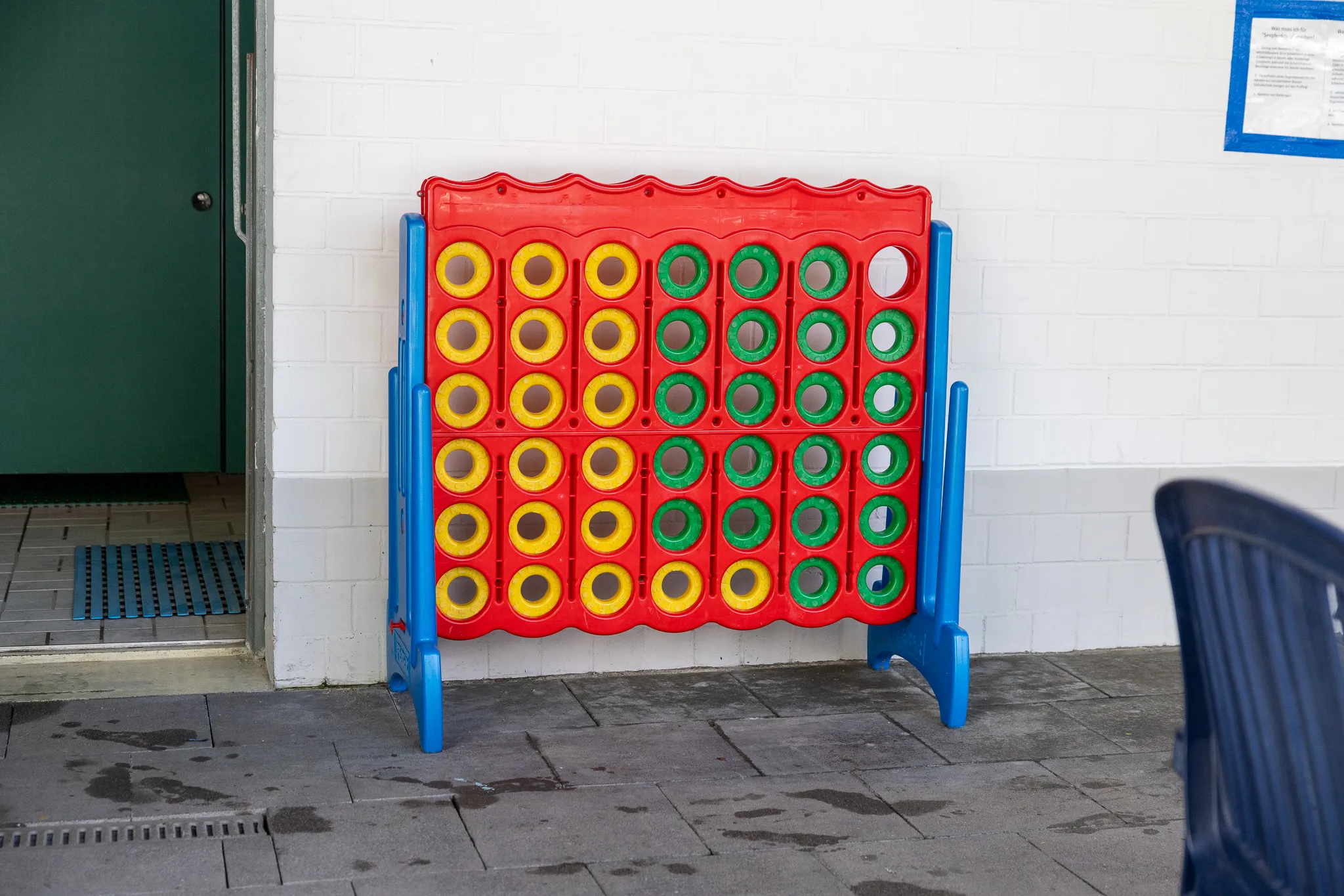 Rotes Vier Gewinnt Spiel steht auf einem grauen Pflaster vor einer weißen Wand und einer grünen Tür.