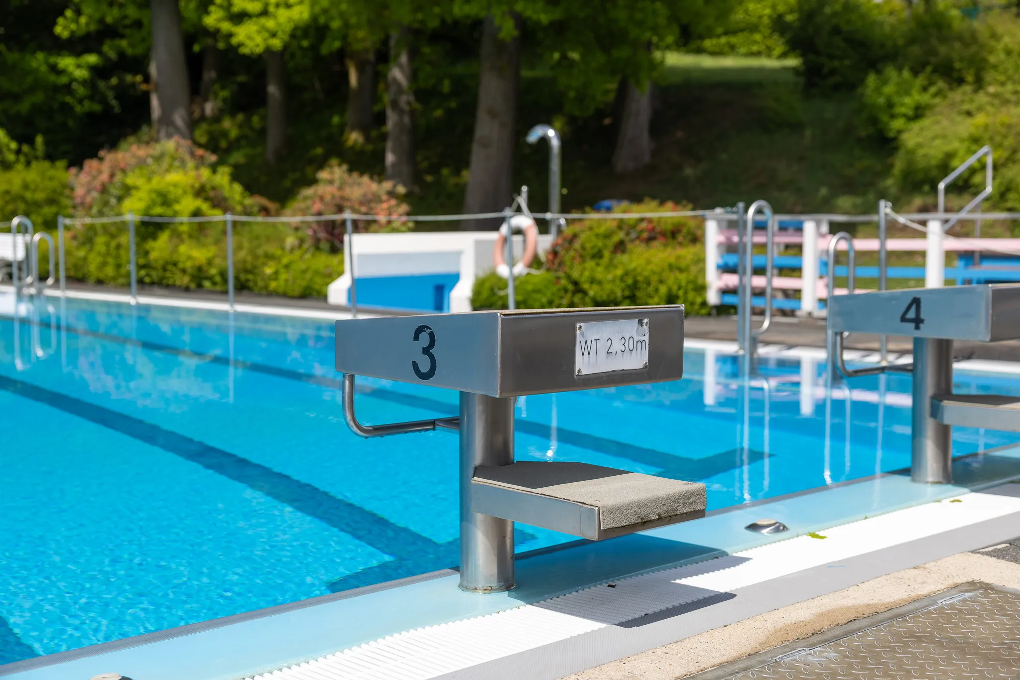 Startblöcke mit den Nummern 3 und 4 im blauen Wasser eines Schwimmbades, im Hintergrund Bäume und Büsche.
