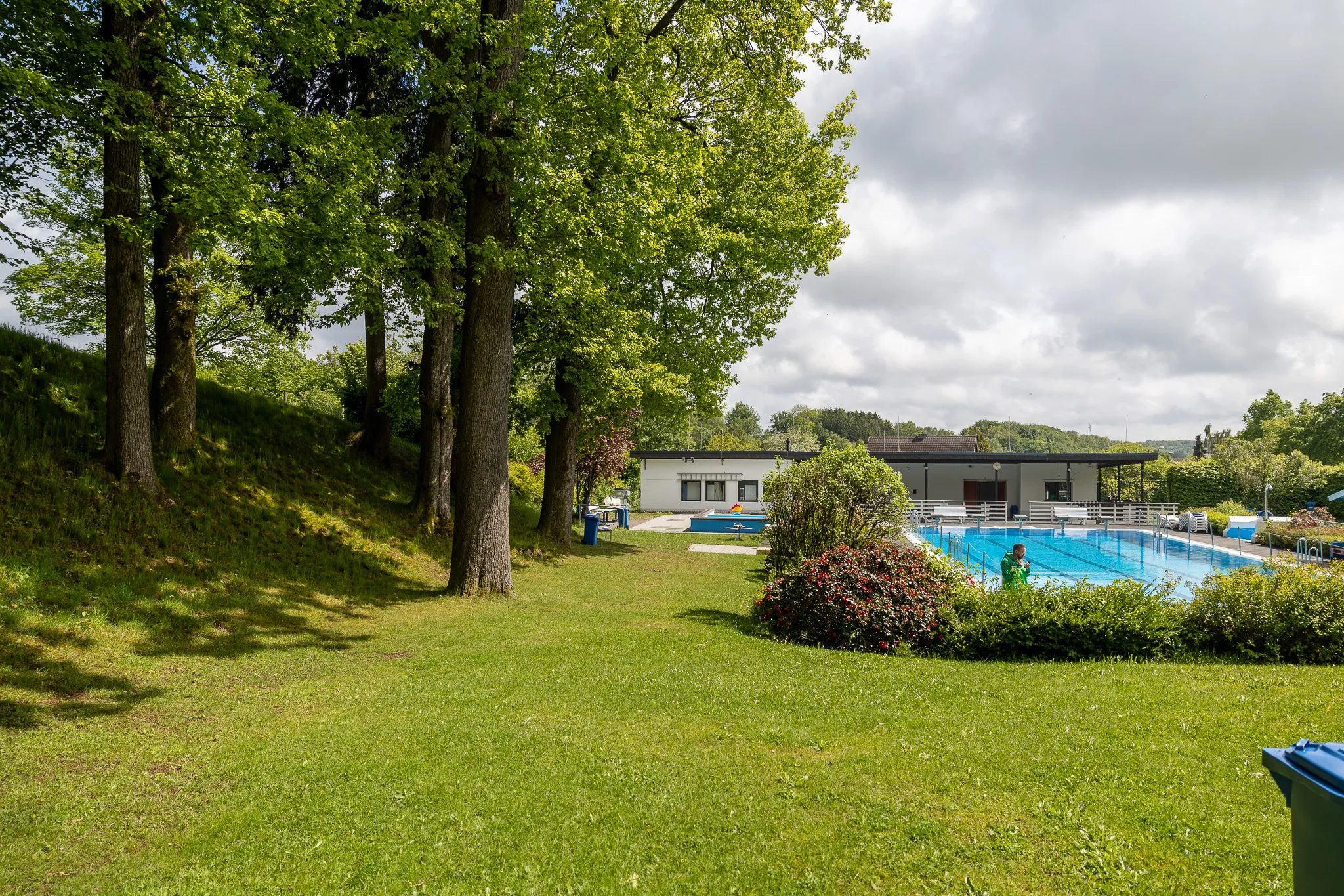 Grüner Rasen vor einem Freibad mit Becken, Liegeflächen und einem Gebäude im Hintergrund.