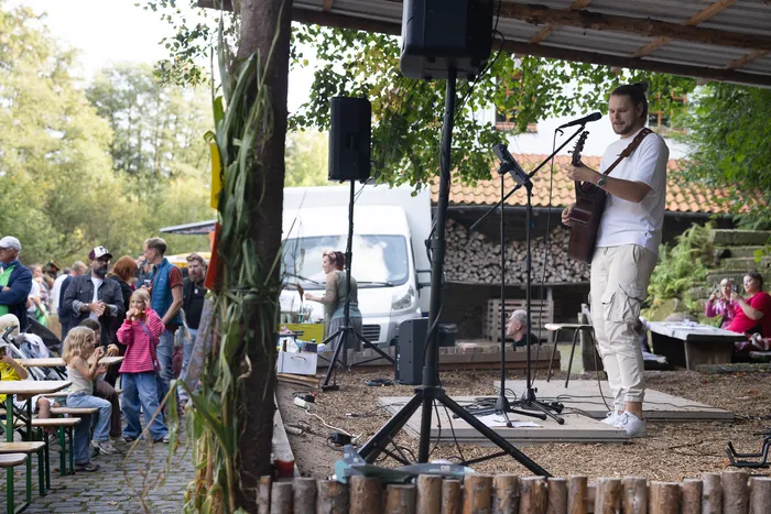 Ein Musiker spielt Gitarre und singt in ein Mikrofon auf einer überdachten Bühne bei einer Veranstaltung im Freien.