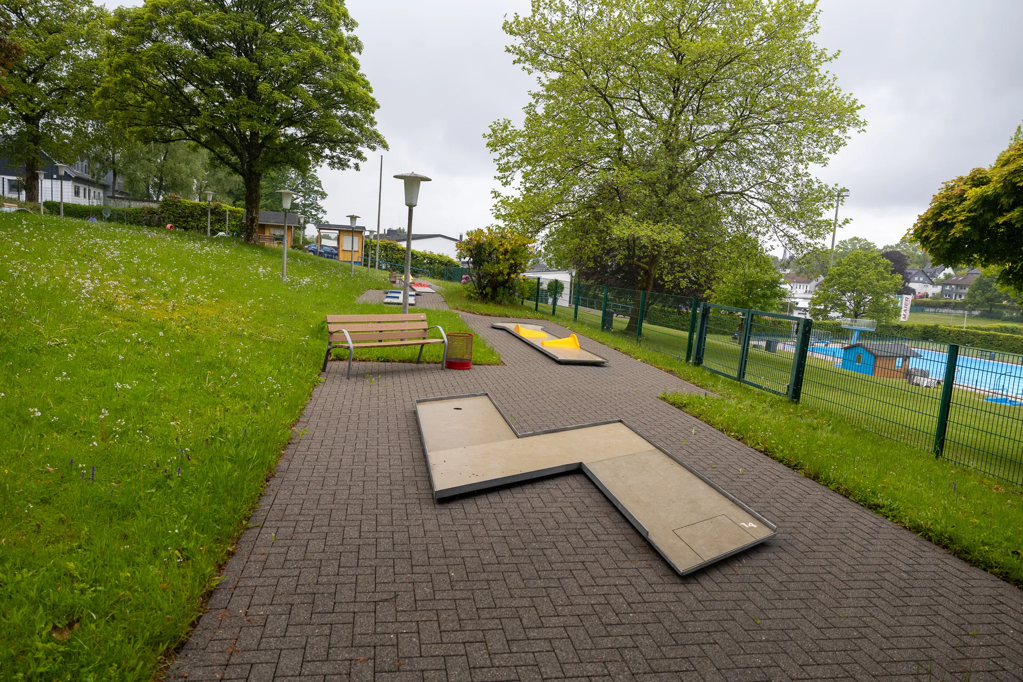 Minigolfanlage im Freien neben einem Freibad mit blauem Wasser und grüner Rasenfläche.