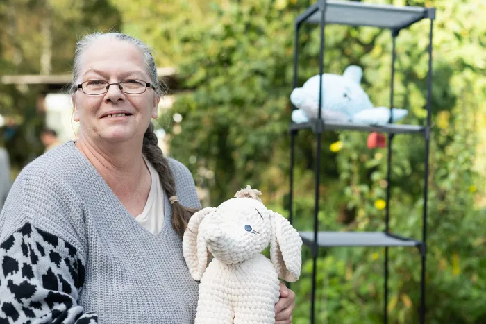 Eine Frau mit Brille hält ein großes, weißes, gehäkeltes Elefanten-Stofftier vor einem Hintergrund aus grünen Pflanzen.