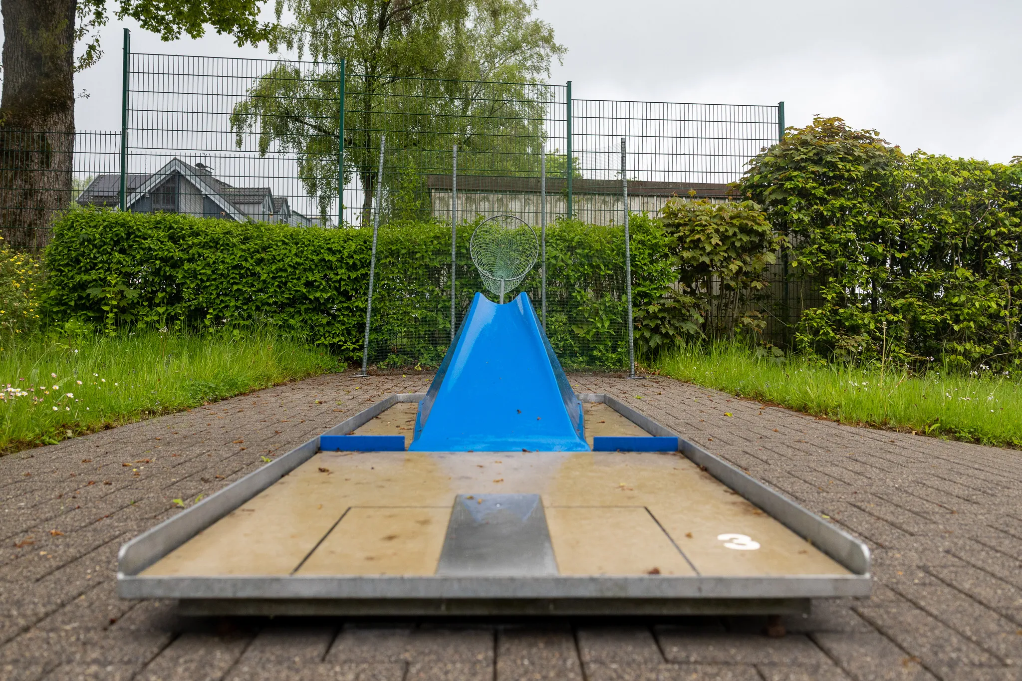 Minigolfbahn mit blauem Hindernis und Loch im Zentrum, umgeben von Rasen und Büschen.