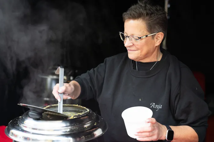 Eine Frau mit Brille schöpft Essen aus einem dampfenden Metallbehälter in einen weißen Becher.