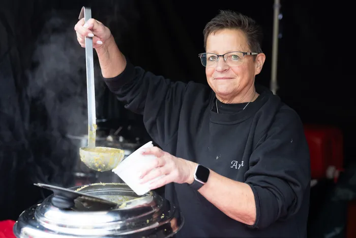 Eine Frau schöpft mit einer Metallkelle dampfendes Essen aus einem silbernen Warmhaltebehälter in einen weißen Becher.