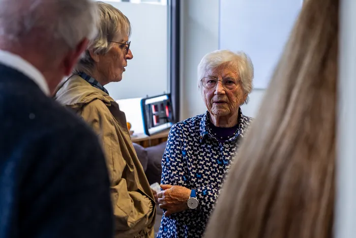Zwei ältere Frauen unterhalten sich in einem Raum, während im Hintergrund weitere Personen zu sehen sind.