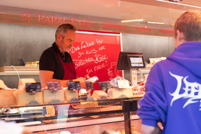 Ein Mann in roter Schürze bedient eine Käsetheke bei „PAPAGENO“. Ein Schild im Hintergrund lautet: „Das Leben ist zu kurz, um schlechten Käse zu essen!“.