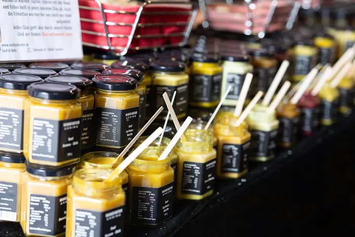 Zahlreiche kleine Gläser mit gelbem Inhalt und schwarzen Etiketten auf einem Marktstand, teilweise mit Holzstäbchen.