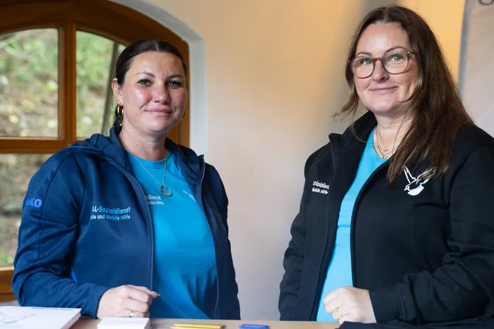 Zwei Frauen in blauen und schwarzen Jacken mit dem Logo des AL-Sozialdienstes blicken in die Kamera.