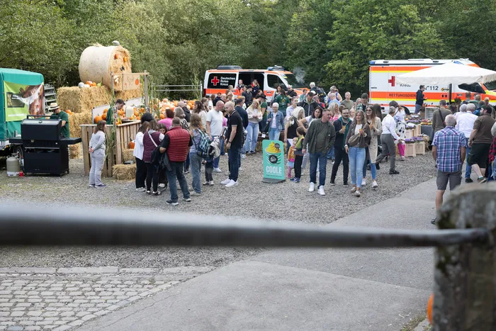 Menschenmenge bei einem herbstlichen Fest im Freien mit Kürbissen auf Heuballen und Rettungsfahrzeugen im Hintergrund.