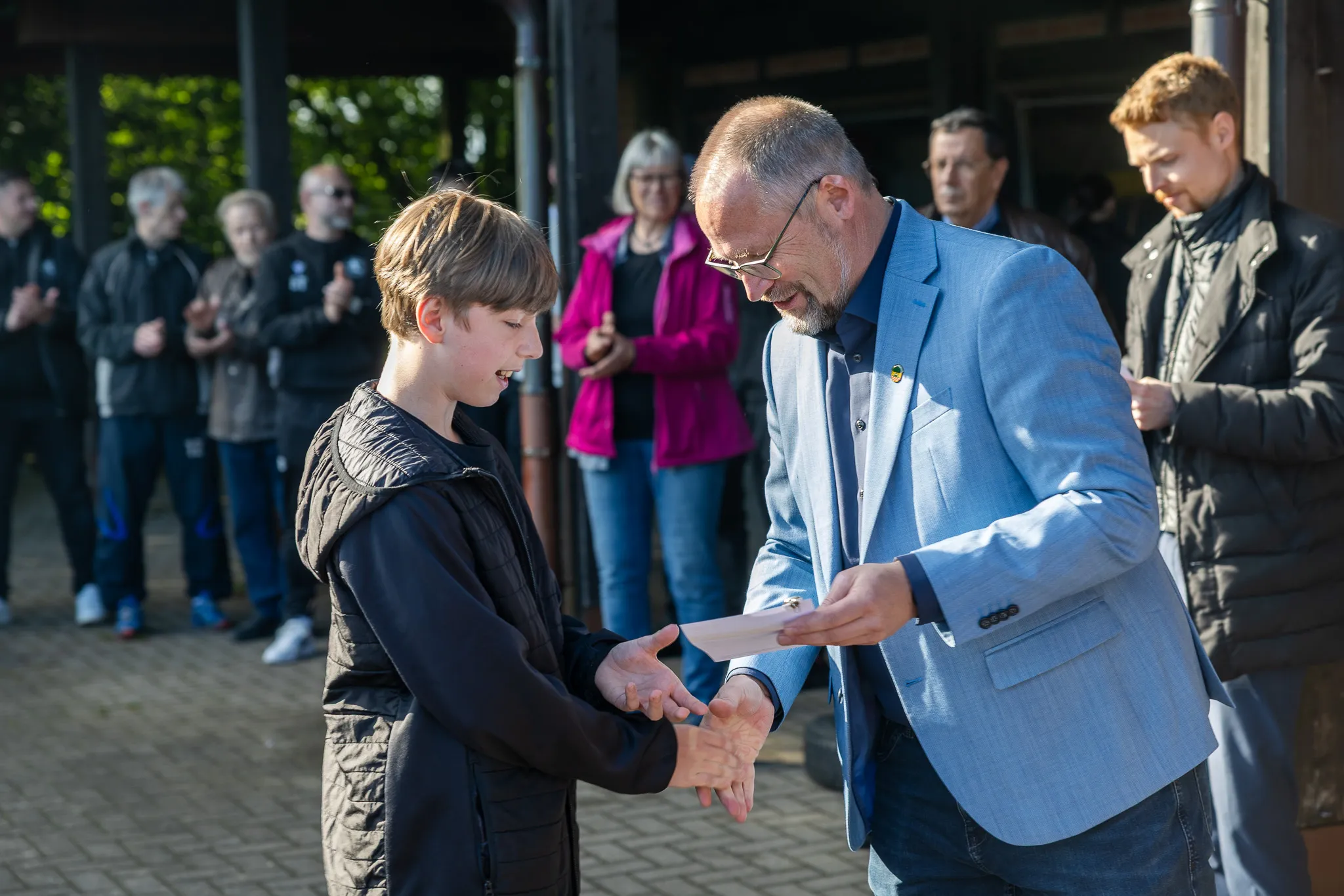 Ein Mann in einem hellblauen Anzug überreicht einem jungen Menschen ein Dokument. Im Hintergrund stehen weitere Personen auf einem Pflasterweg.