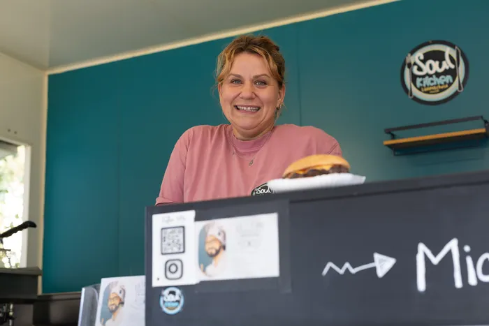 Eine lächelnde Frau steht hinter einem Verkaufstresen mit einem Burger vor einer blaugrünen Wand mit dem „Soul Kitchen“-Logo.