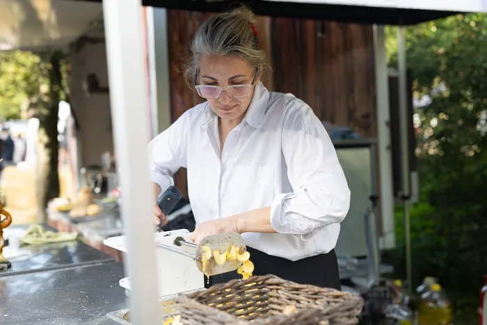 Eine Frau in weißer Kleidung schneidet mit einer Mandoline gelbe Gemüsestücke in einen Weidenkorb an einem Verkaufsstand.