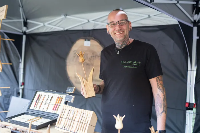 Ein lächelnder Mann mit Tattoos präsentiert eine geschnitzte Holzblume an einem Marktstand unter einem Zelt.