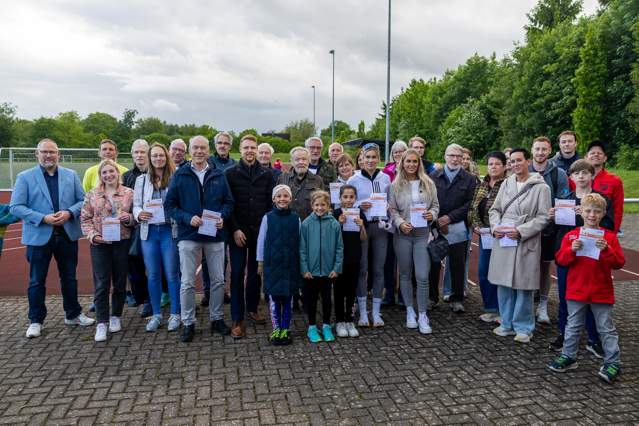 Eine Gruppe von etwa dreißig Menschen steht lächelnd auf einem gepflasterten Platz vor einem Tennisplatz und hält Urkunden in den Händen.
