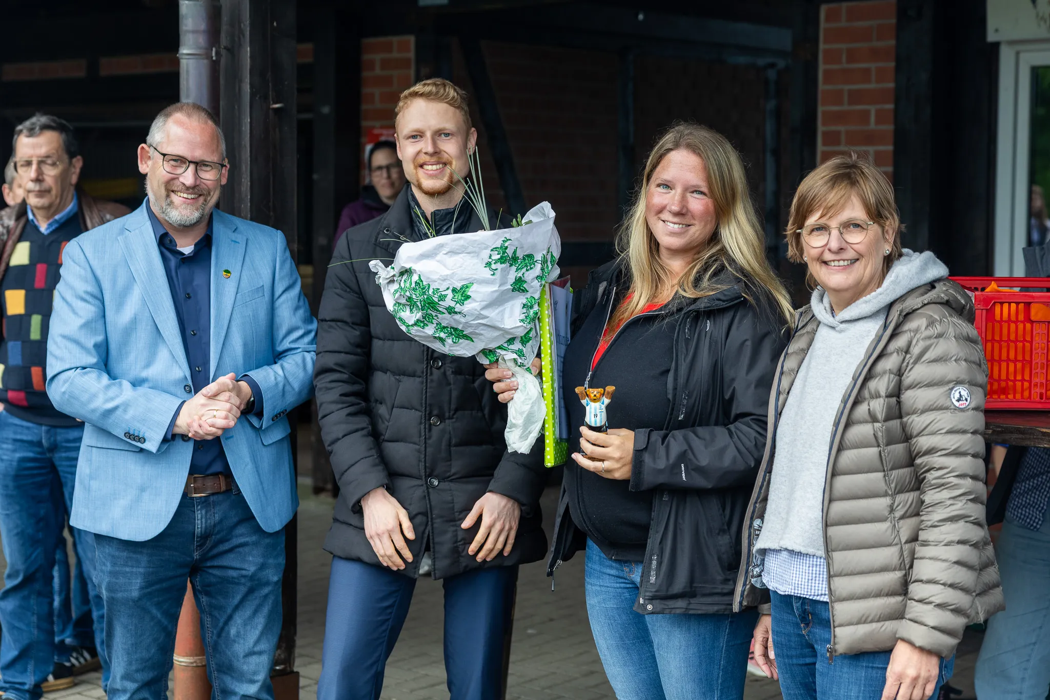 Vier lächelnde Menschen stehen im Freien vor einem Gebäude. Ein Mann hält einen Blumenstrauß.