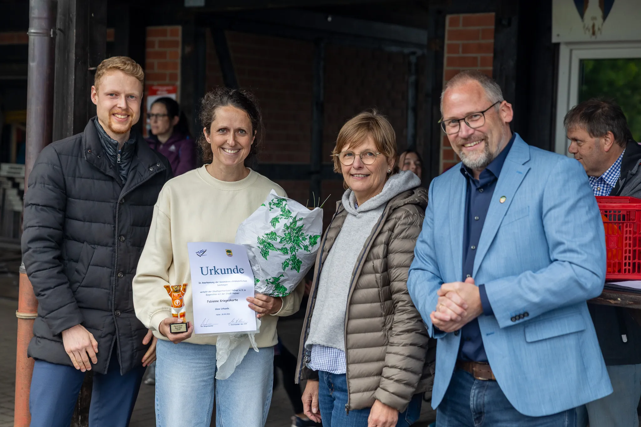 Fabienne Kriegeskorte nimmt eine Urkunde und einen Blumenstrauß von zwei Männern und einer Frau entgegen, während ein weiterer Mann lächelt.
