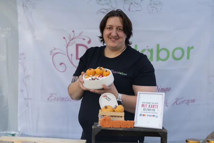 Eine lächelnde Frau hält eine weiße Schale mit kleinen Zierkürbissen vor einem Banner der Marke Dekolebor.