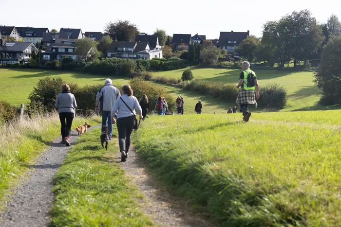 Eine Gruppe von Menschen wandert mit Hunden auf einem Feldweg durch eine grüne Wiese, im Hintergrund stehen Wohnhäuser.