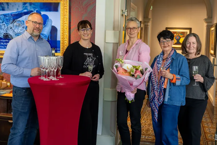 Vier Personen stehen vor einem Tisch mit Weingläsern und einem Kunstwerk im Hintergrund. Zwei Frauen halten Blumensträuße.