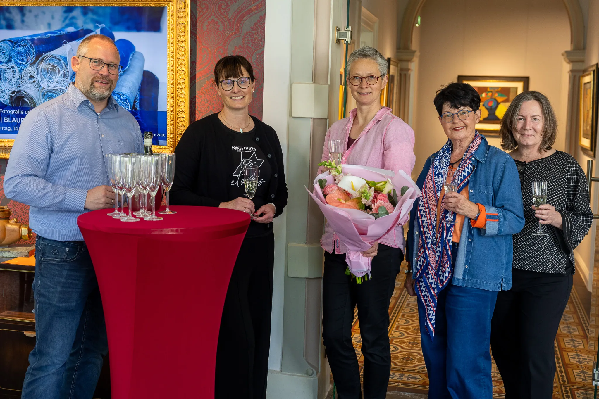 Vier Personen stehen vor einem Tisch mit Weingläsern und einem Kunstwerk im Hintergrund. Zwei Frauen halten Blumensträuße.