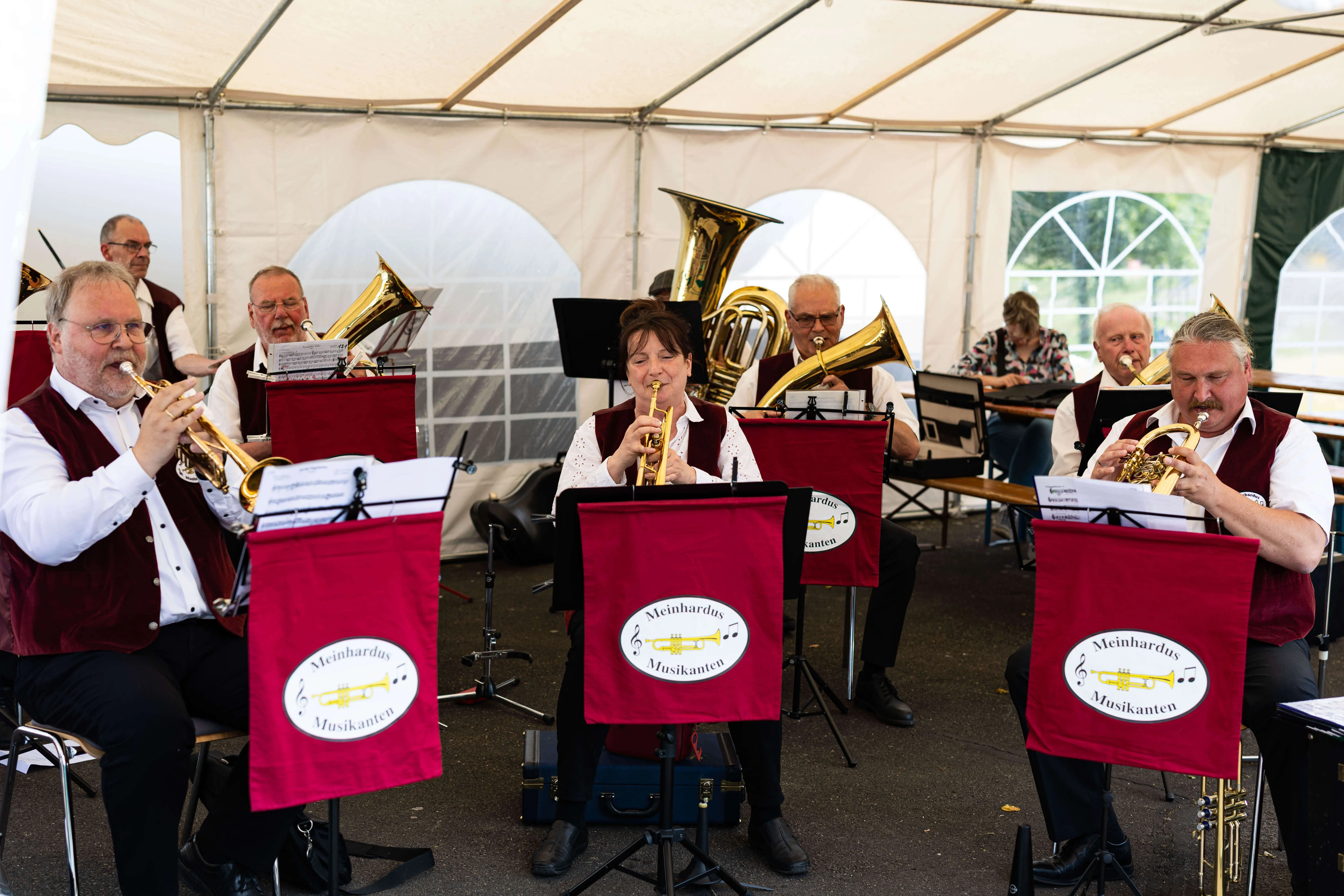 Blasmusikensemble spielt unter einem weißen Zelt. Die Musikerinnen und Musiker sitzen auf Stühlen vor Notenständern und spielen verschiedene Blechblasinstrumente.