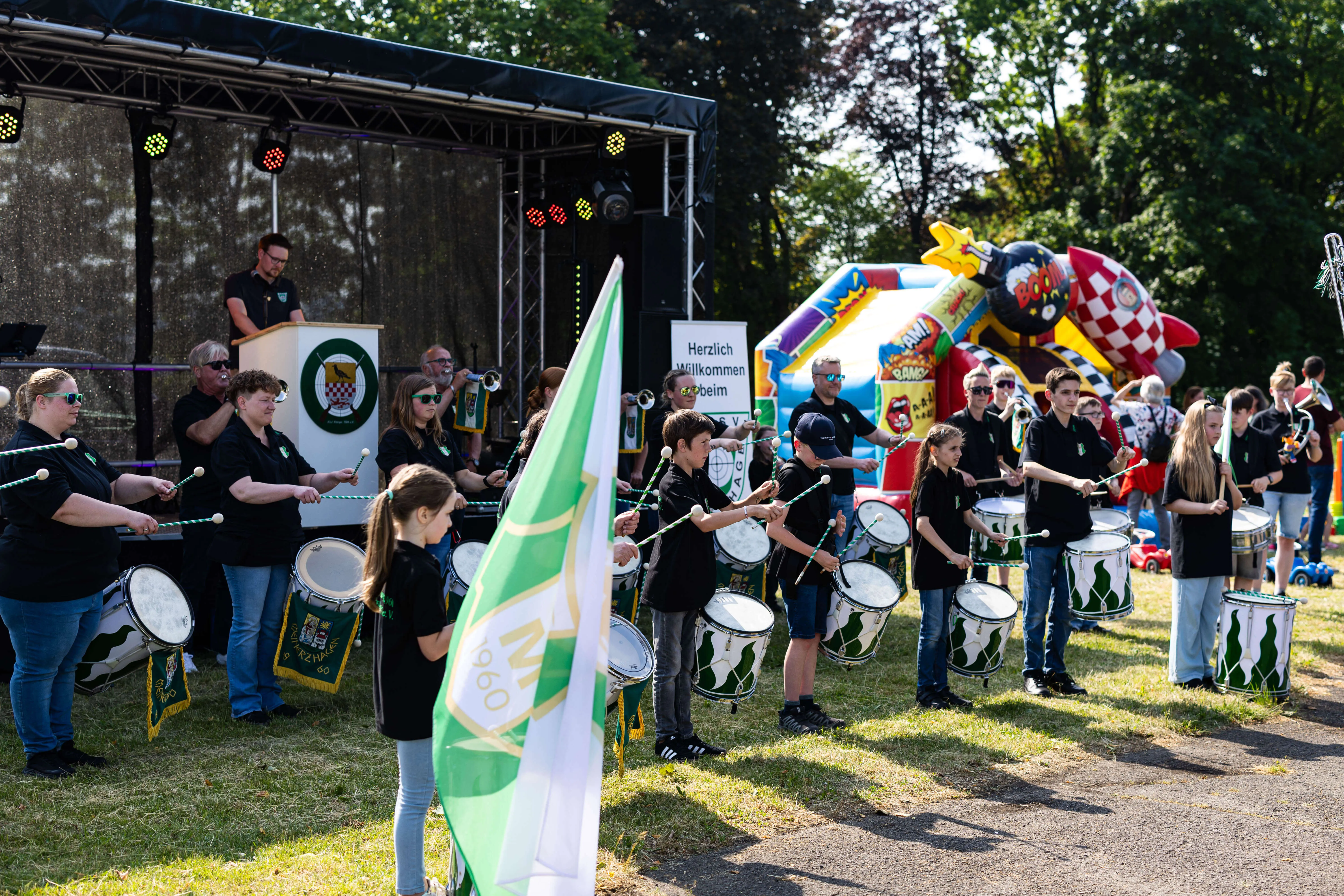 Musikzug des KKSV Meinerzhagen spielt auf dem Schützenplatz während des Jubiläumsfests. Im Hintergrund eine Hüpfburg und eine Bühne mit der Aufschrift „Herzlich Willkommen beim“.