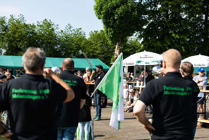 Mehrere Personen stehen auf einem Festplatz mit grünen und weißen Fahnen, während ein Mann im Hintergrund Trompete spielt. Im Hintergrund sind Biertischgarnituren und Schirme zu sehen.