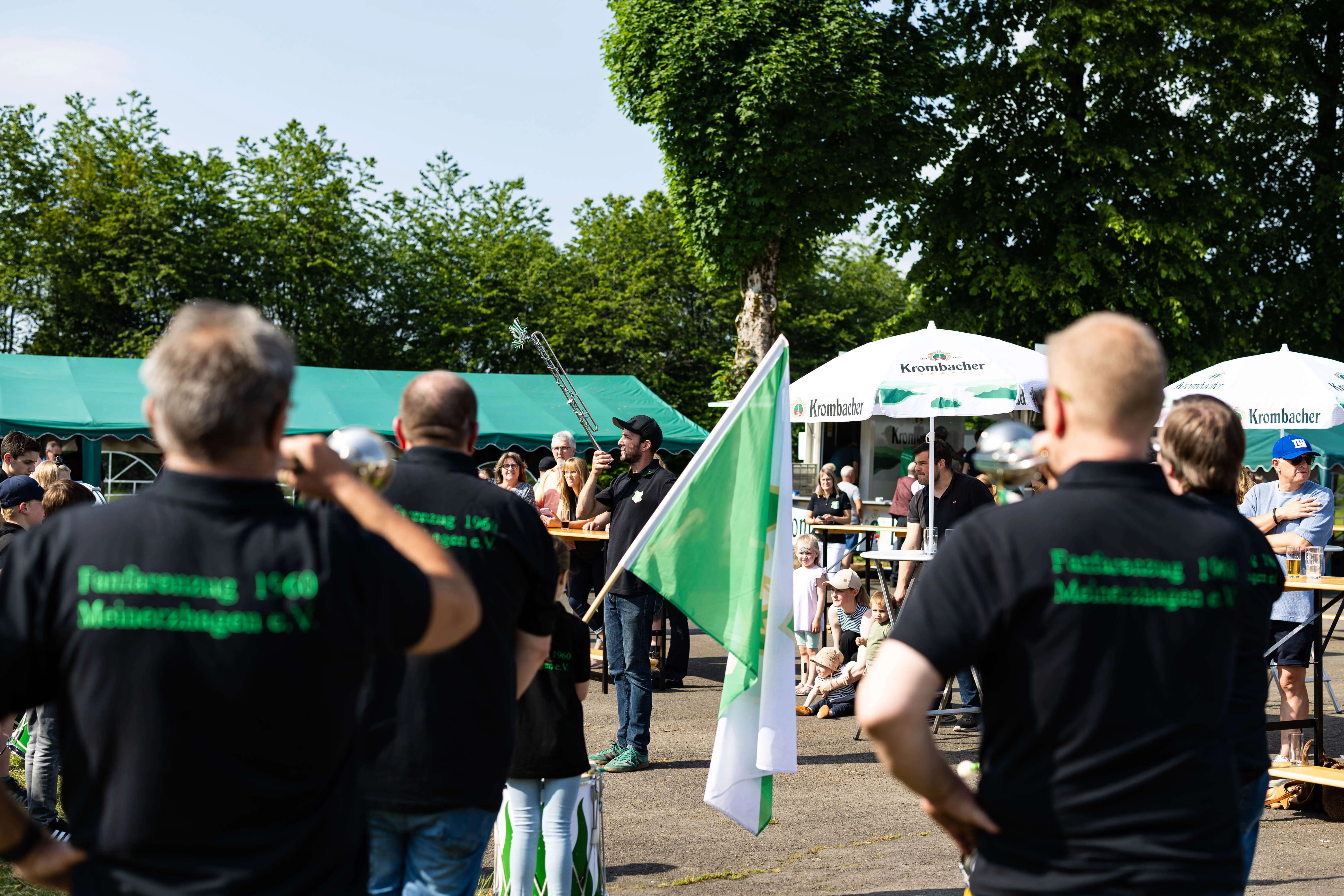 Mehrere Personen stehen auf einem Festplatz mit grünen und weißen Fahnen, während ein Mann im Hintergrund Trompete spielt. Im Hintergrund sind Biertischgarnituren und Schirme zu sehen.