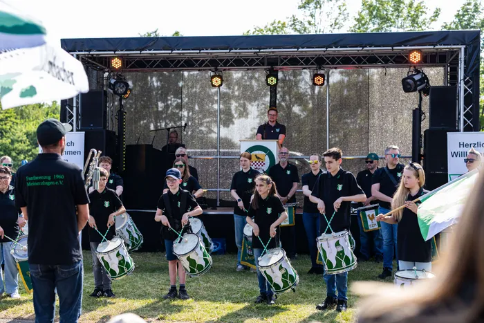 Eine Gruppe von Kindern und Jugendlichen spielt auf einer Bühne Trommeln, während ein Mann im Hintergrund dirigiert. Im Hintergrund sind Banner und Bühnenbeleuchtung zu sehen.
