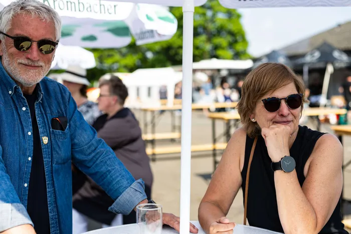 Zwei lächelnde Menschen sitzen an einem Tisch im Freien, im Hintergrund sind weitere Personen und Festzeltplanen zu sehen.