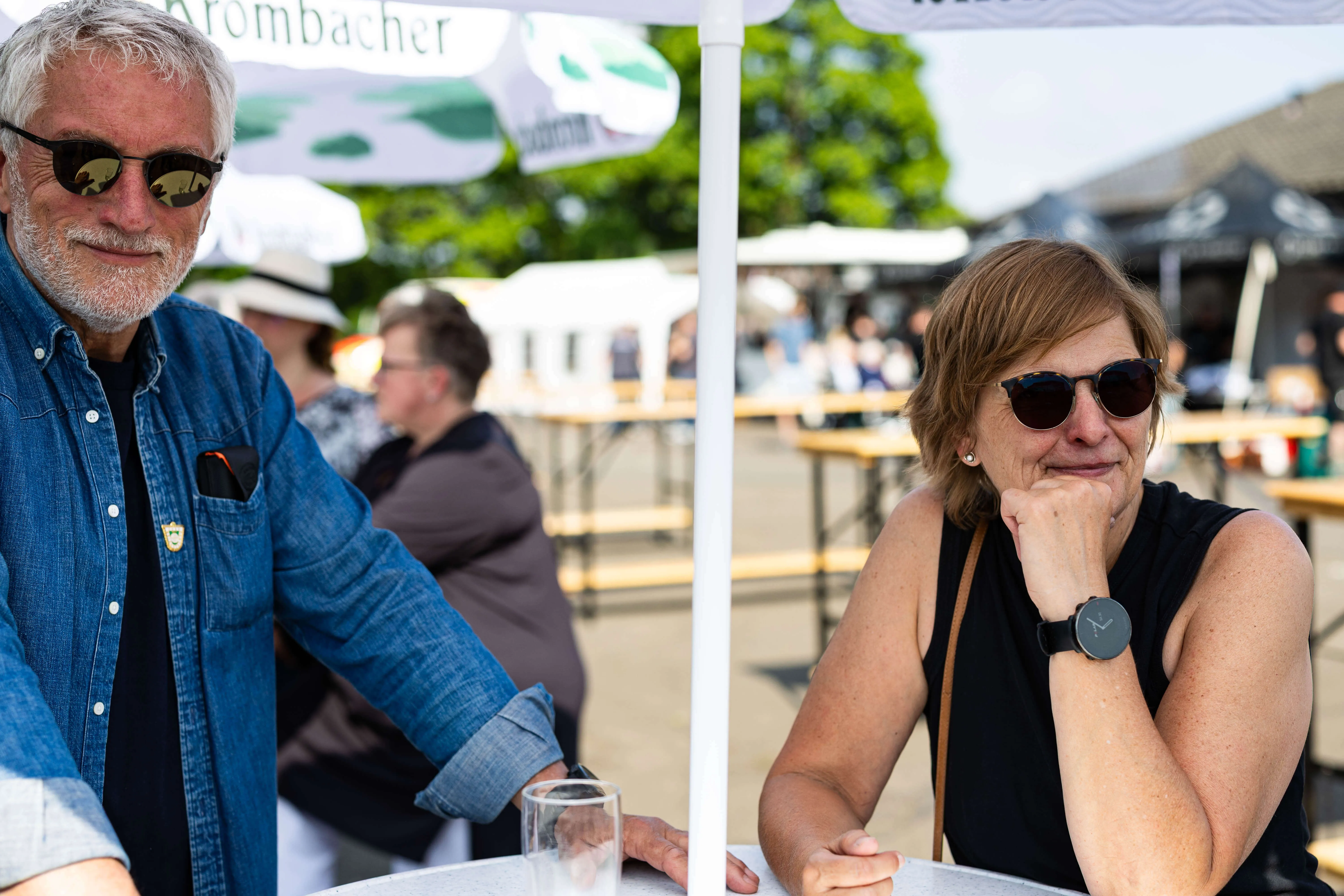 Zwei lächelnde Menschen sitzen an einem Tisch im Freien, im Hintergrund sind weitere Personen und Festzeltplanen zu sehen.