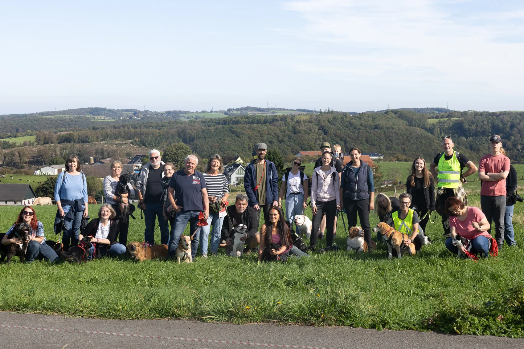 Eine Gruppe von Menschen mit vielen Hunden posiert für ein Gruppenfoto auf einer Wiese vor einer hügeligen Landschaft.