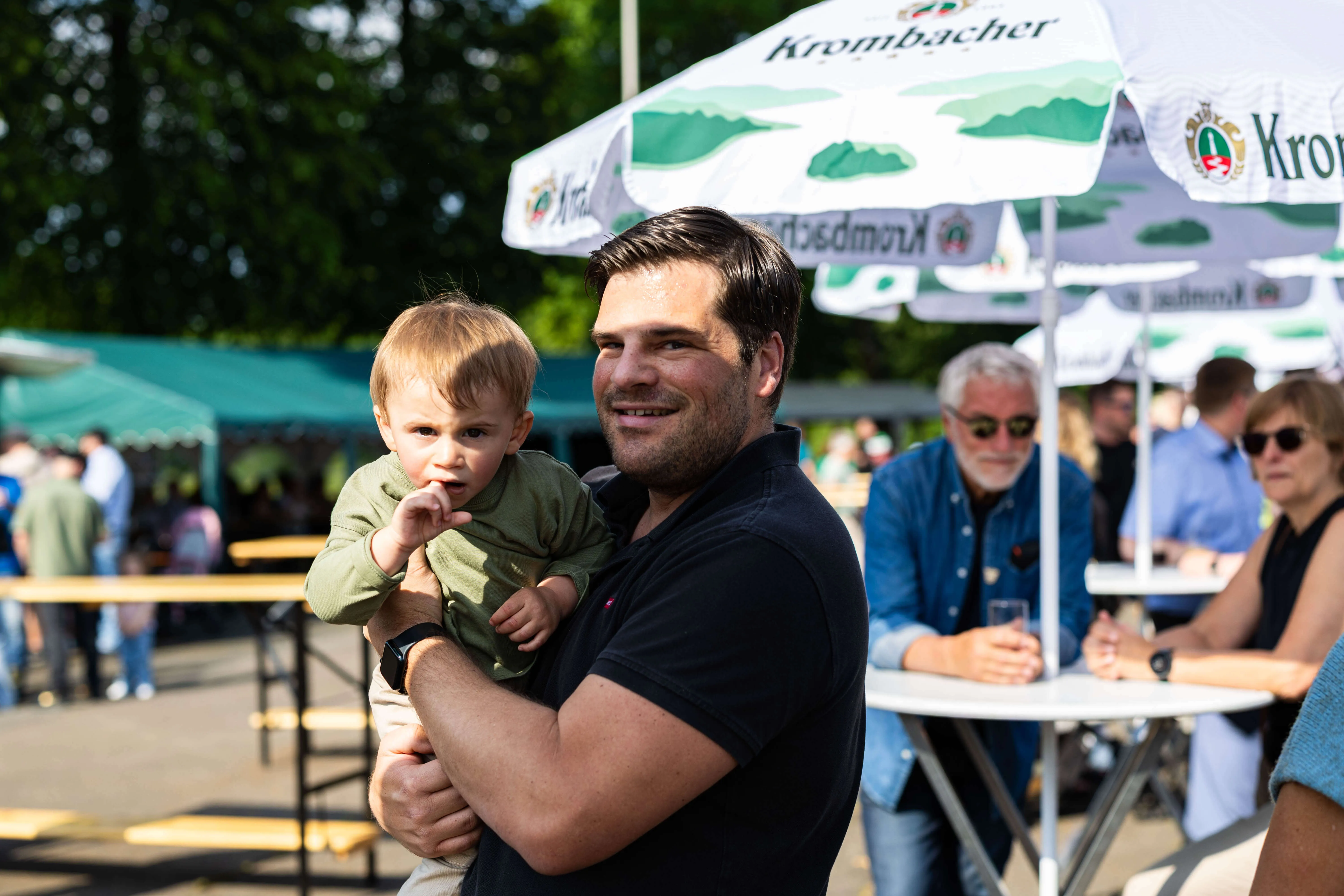 Ein lächelnder Mann hält sein kleines Kind im Arm, im Hintergrund ein Festzelt mit dem Schriftzug „Krombacher“ und weitere Menschen.
