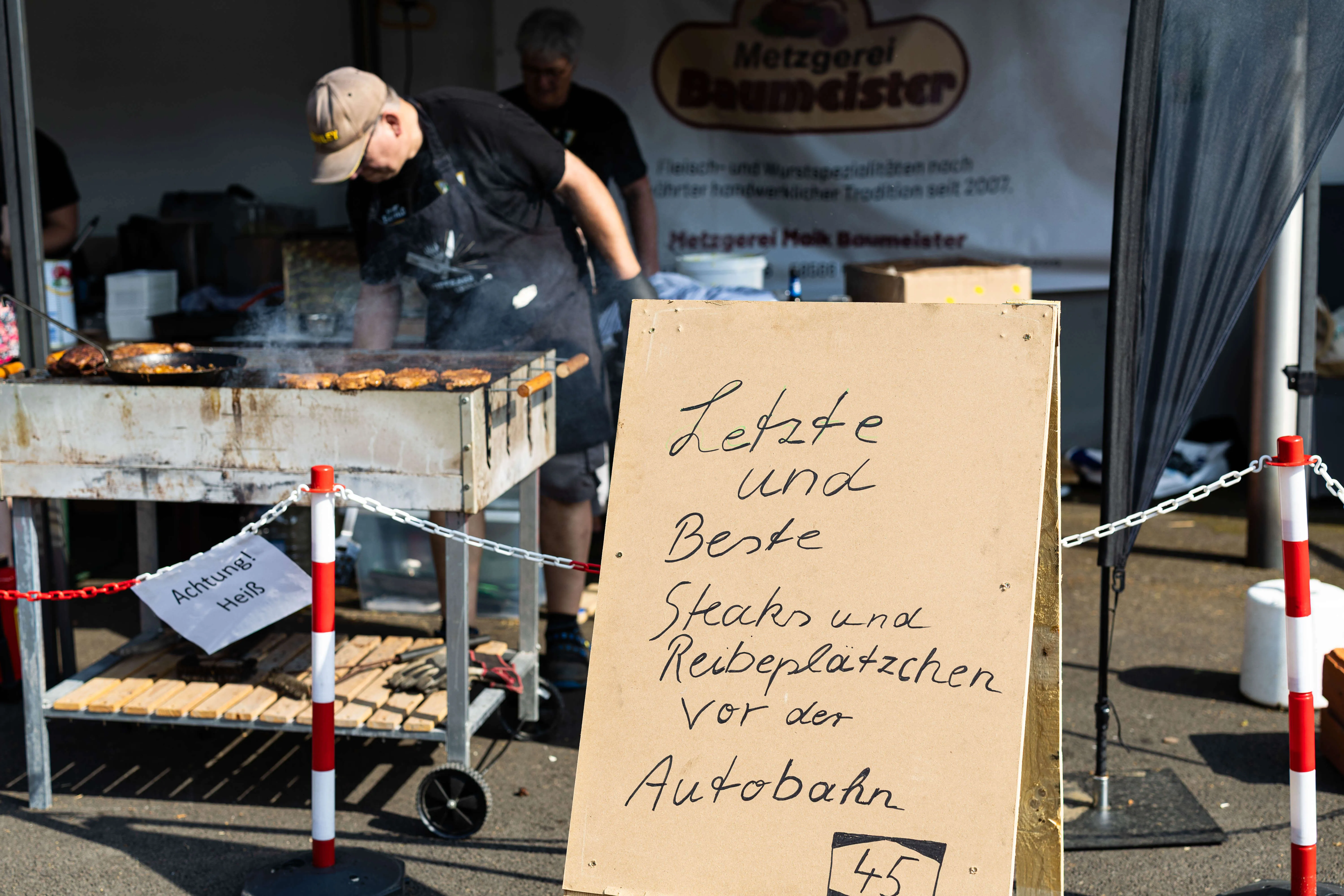 Grill mit Fleisch auf dem Schützenplatz, davor ein Schild mit der Aufschrift „Letzte und Beste Steaks und Reibeküchchen vor der Autobahn“.