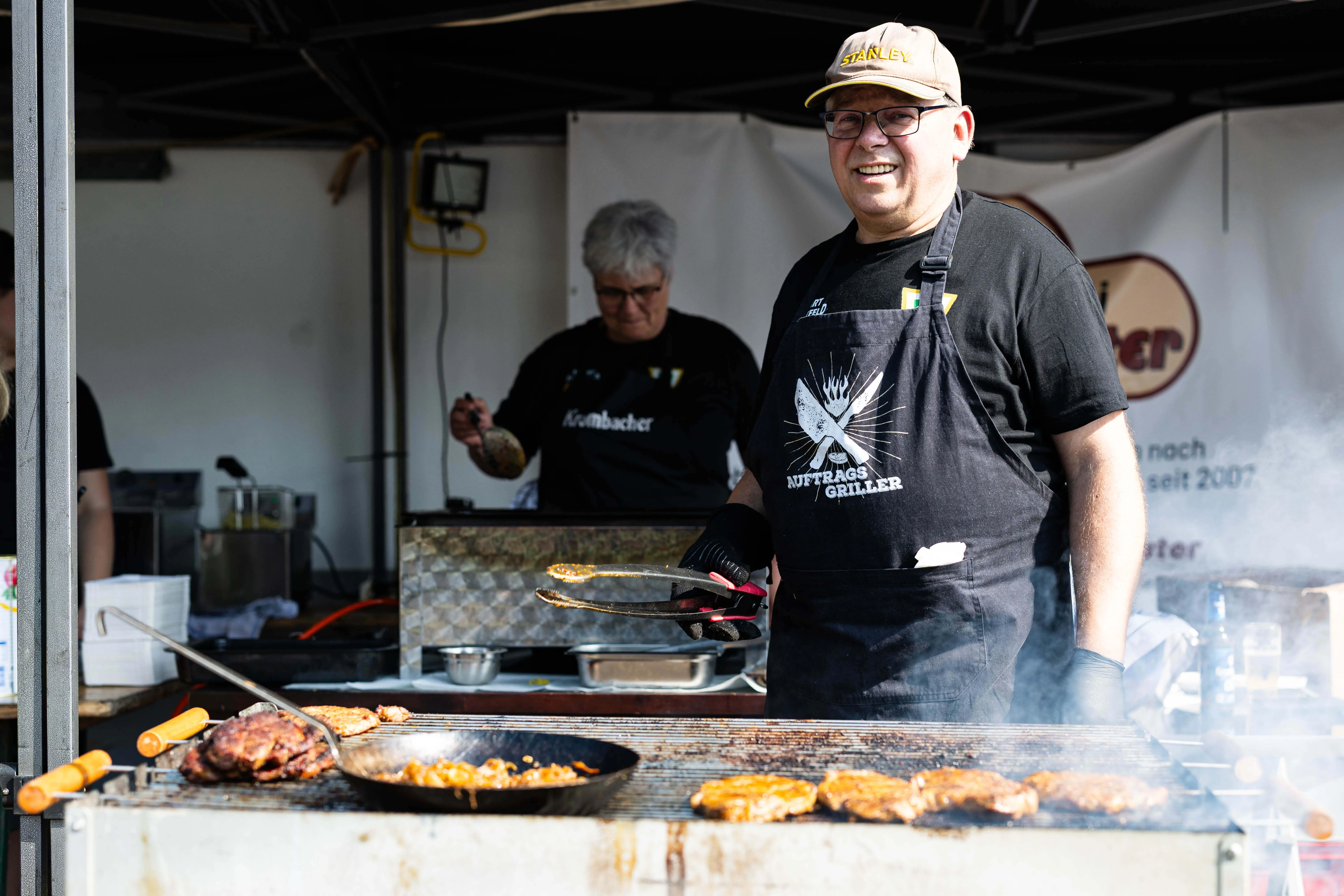 Mann mit Schürze und Kappe steht lächelnd vor einem Grill mit Fleisch, im Hintergrund eine Frau am Stand.
