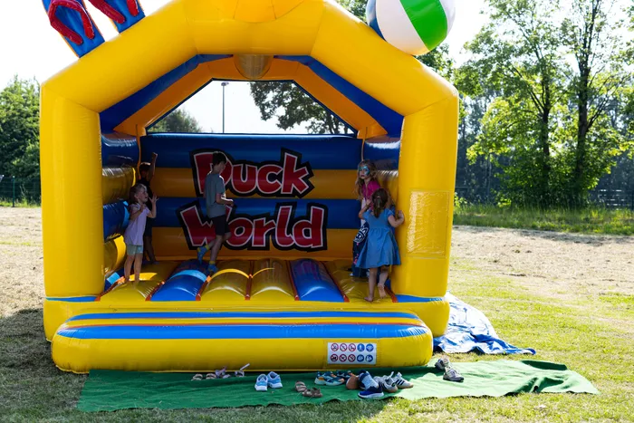 Drei Kinder spielen auf einer gelb-blauen Hüpfburg mit der Aufschrift „Puck World“. Im Vordergrund stehen Schuhe auf einer grünen Matte.