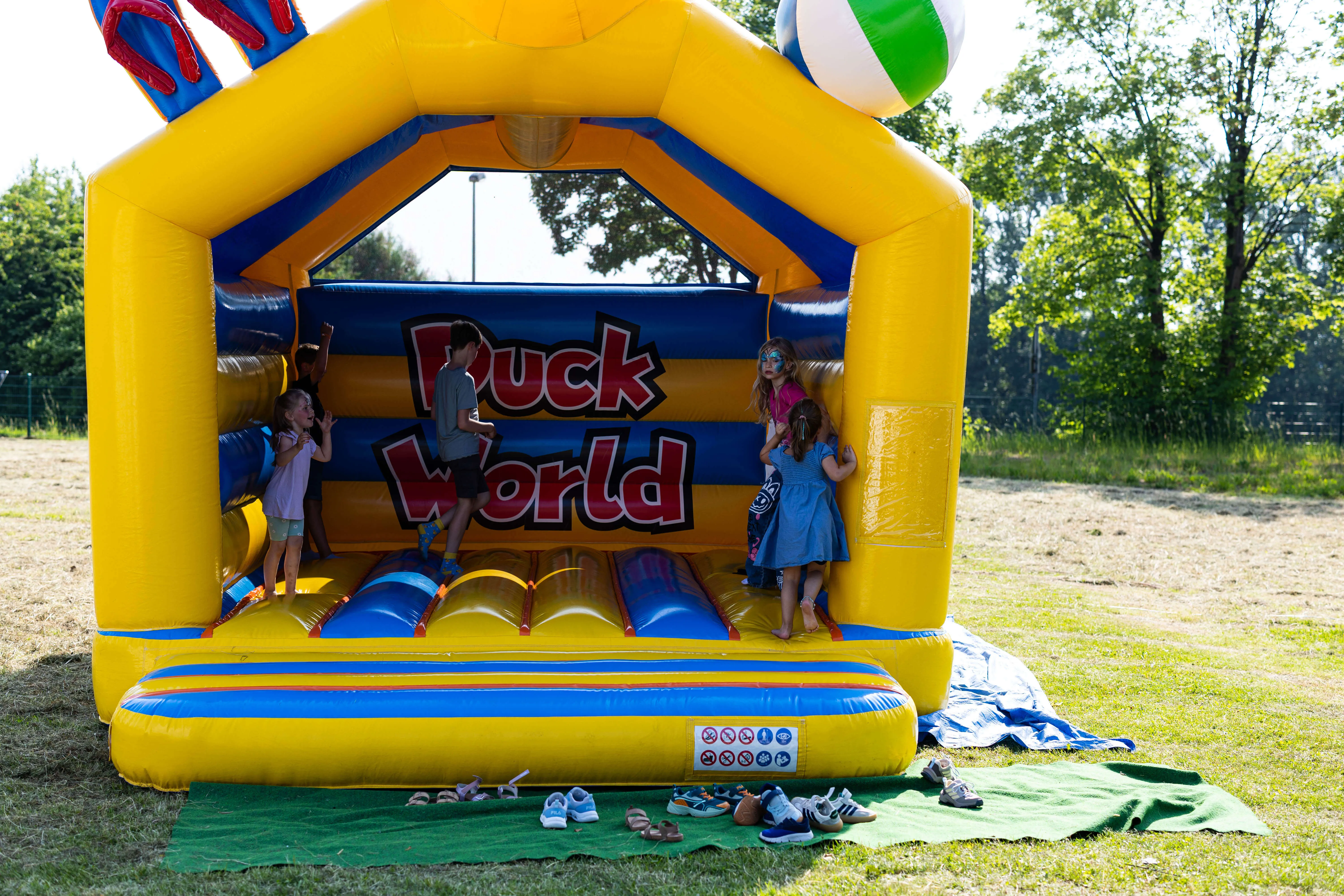 Drei Kinder spielen auf einer gelb-blauen Hüpfburg mit der Aufschrift „Puck World“. Im Vordergrund stehen Schuhe auf einer grünen Matte.