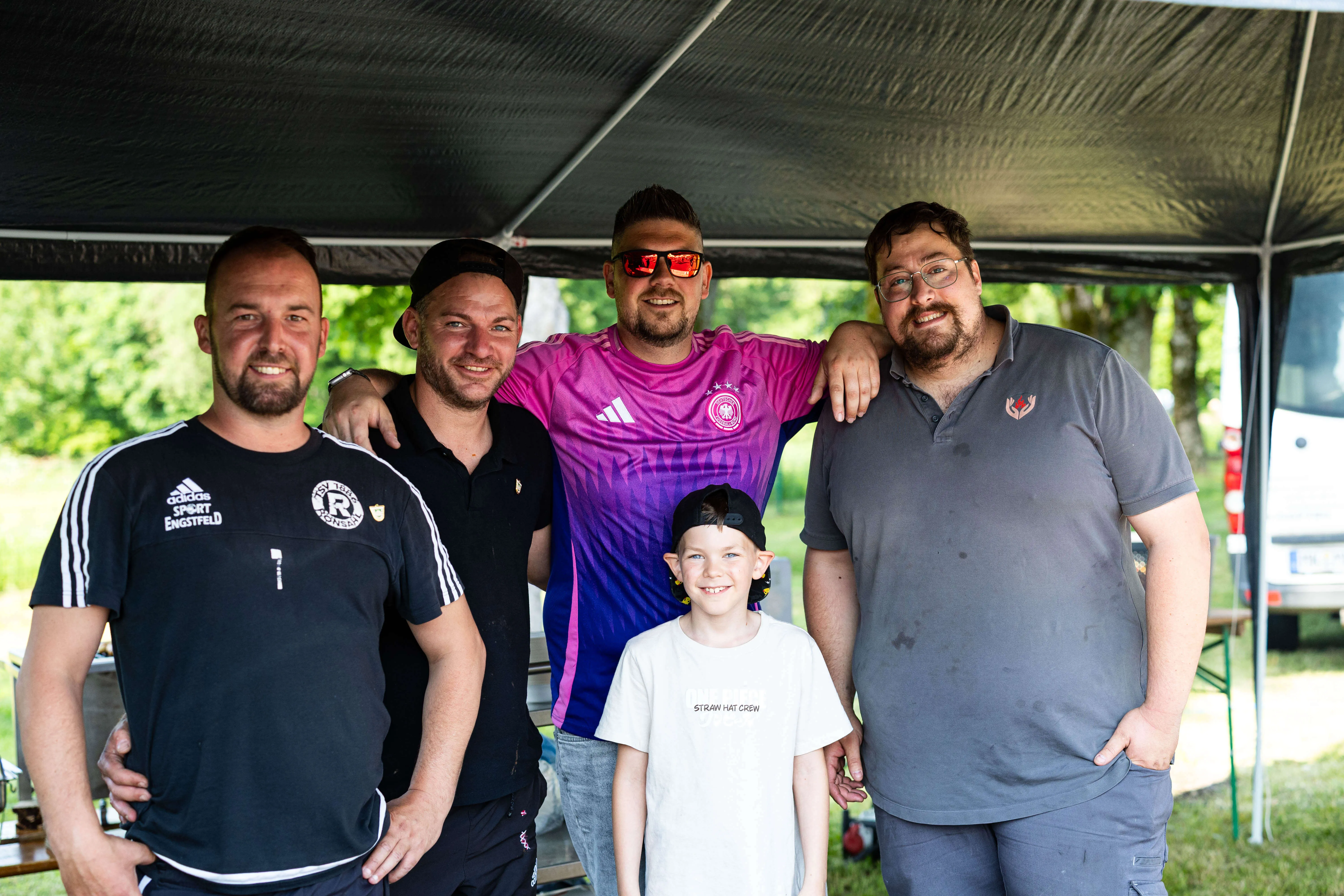 Fünf Männer und ein Junge posieren lächelnd für ein Foto unter einem Zelt. Sie tragen Freizeitkleidung, einige mit Vereinslogos.